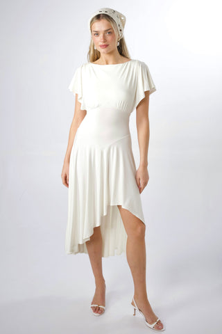 A woman modeling a cream-colored high-low dress with flutter sleeves and a matching patterned headscarf, standing against a light gray studio background.
