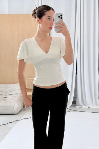 A woman taking a mirror selfie wearing a fitted off-white short-sleeve wrap-style top and black pants in a bright indoor room with white curtains and a light wood wall.