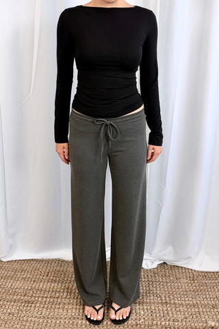 A person modeling wide-leg charcoal gray drawstring sweatpants with a black long-sleeve top, standing against a white draped curtain backdrop.
