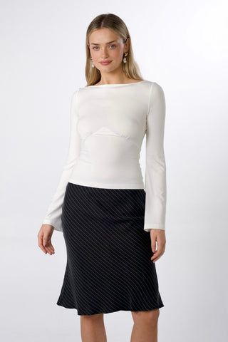 A woman modeling a white long-sleeve boat-neck top and a black knee-length skirt with a subtle diagonal stripe pattern, paired with black thong sandals, against a light gray studio background.