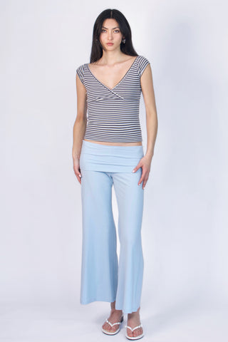 A woman modeling a sleeveless wrap-style top with thin horizontal navy and white stripes, paired with light blue wide-leg pants and white sandals, against a plain light gray studio background.