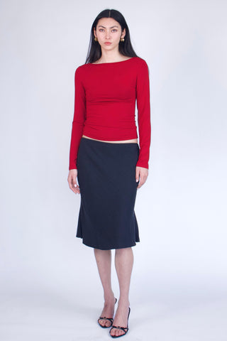 A model stands against a light gray studio background wearing a bright red long-sleeve boat-neck top and a dark navy midi skirt, paired with black strappy heels.
