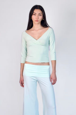 A model wearing a pale mint green wrap-style three-quarter sleeve crop top and matching light blue wide-leg pants, standing against a light gray studio background.
