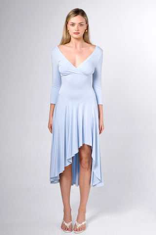 A woman modeling a light blue long-sleeve high-low dress with a V-neckline and draped front detail, photographed against a plain light gray studio background.