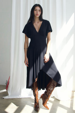 Woman wearing a black dress and brown boots standing in front of white curtains.