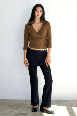 Woman wearing a brown top and black pants against a white wall